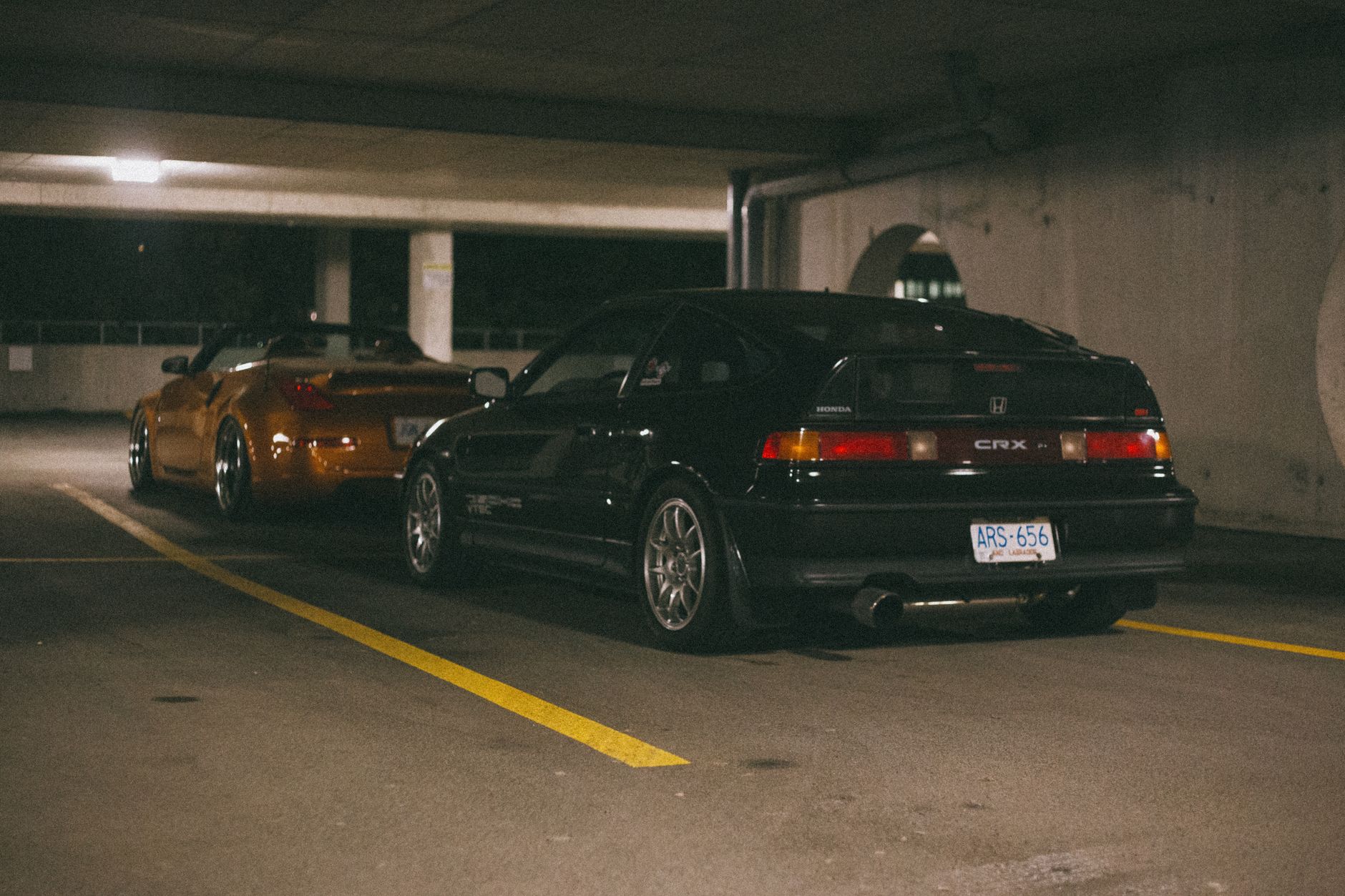 Black Honda CR-X and gold sports car parked in dimly lit garage.