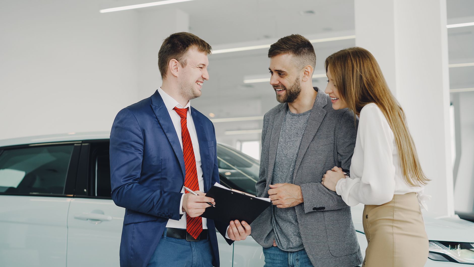 A smiling couple buys a new car from a confident salesman inside a modern car dealership.