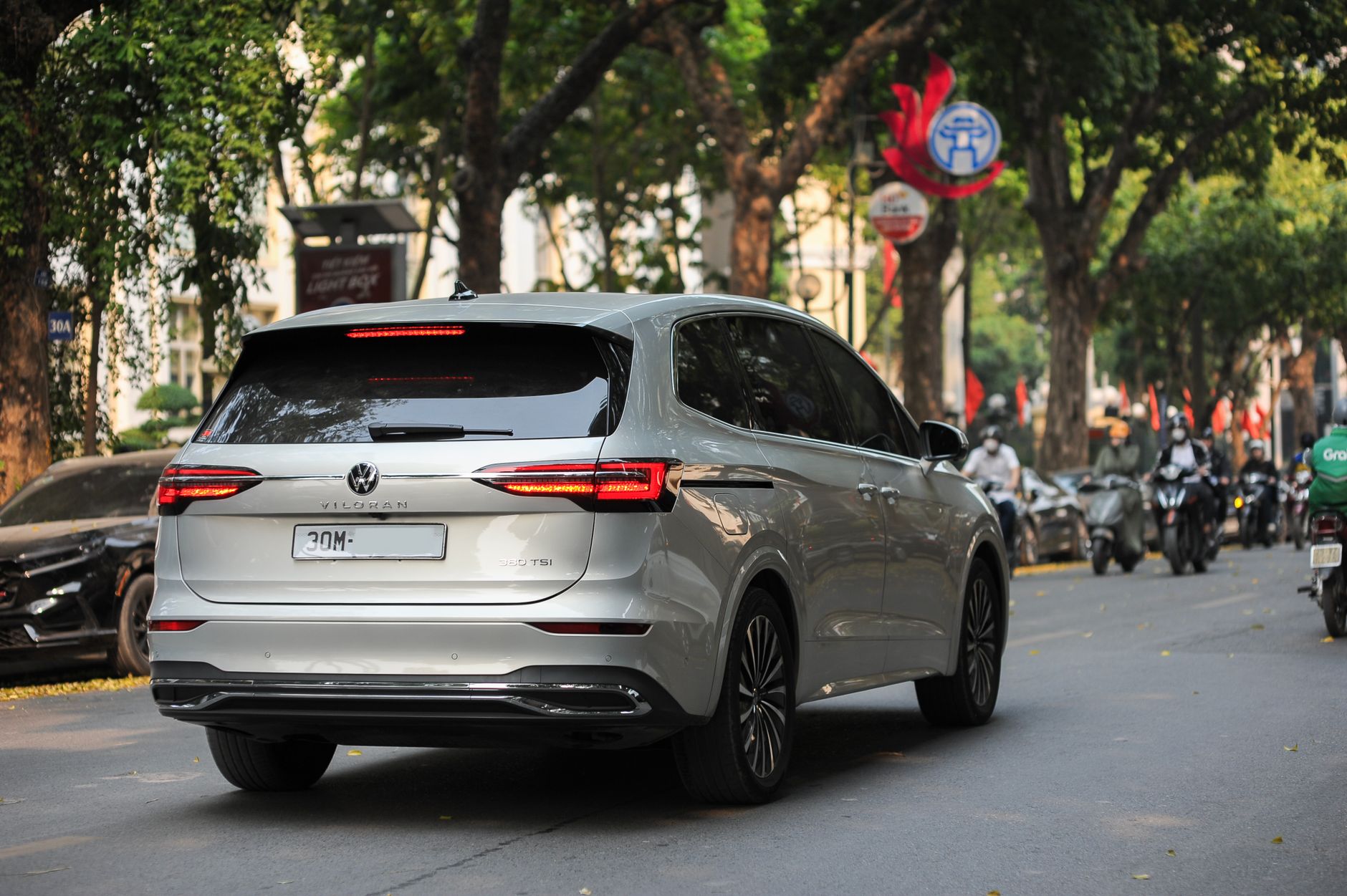 Modern silver MPV driving through a leafy urban street with a busy city backdrop.