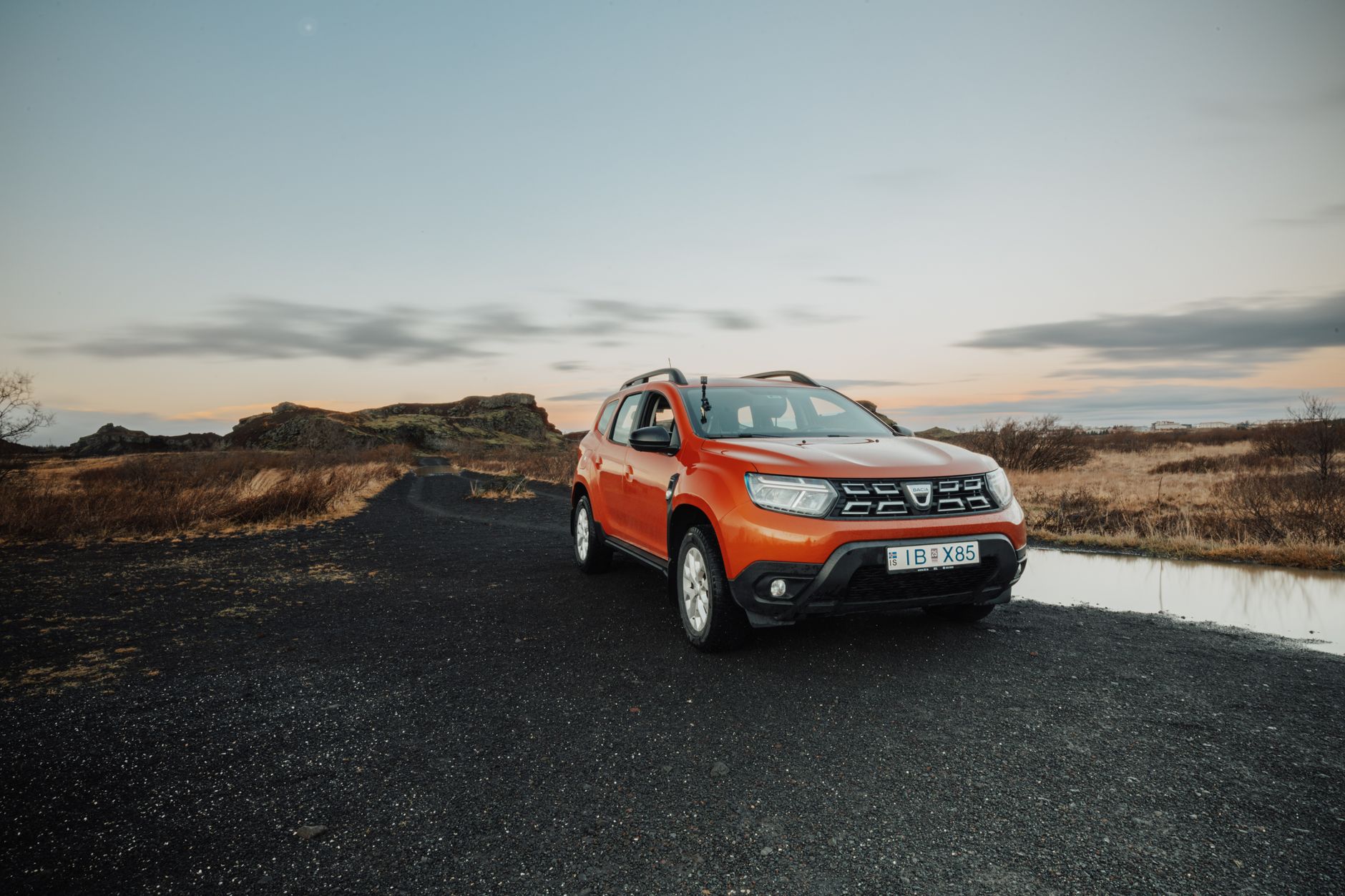 Orange Dacia Duster SUV parked on a rural dirt road during a beautiful sunset, featuring vast landscapes.