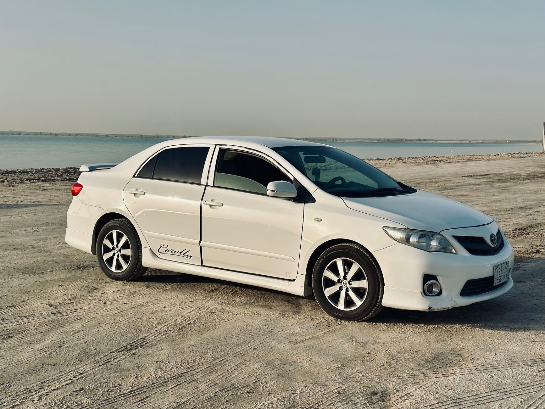 A pristine white Toyota Corolla parked on a sandy beach with a serene ocean backdrop.