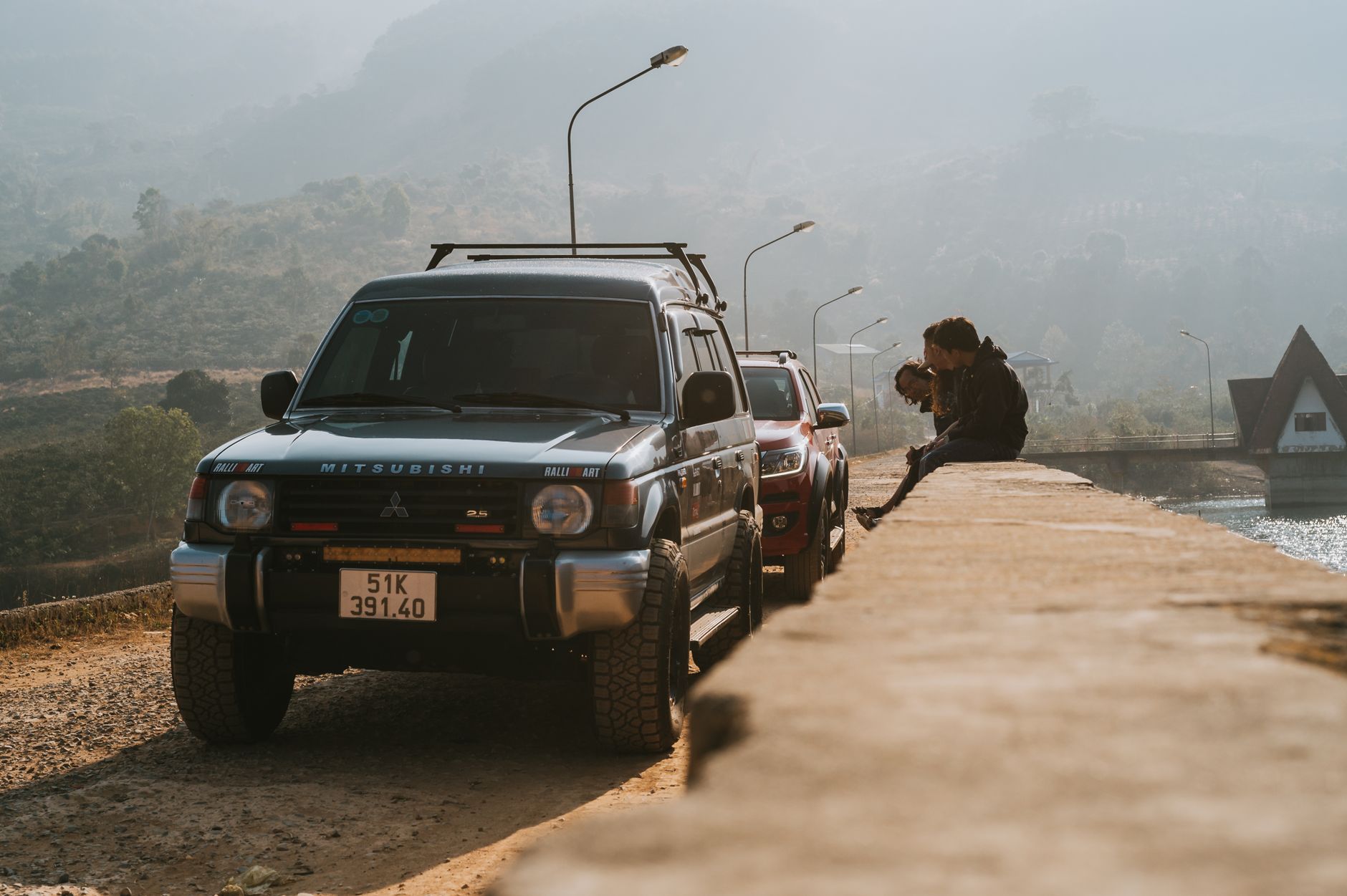 A Mitsubishi SUV and cars parked on a mountain bridge during sunrise with hills in the backdrop and hazy atmosphere.