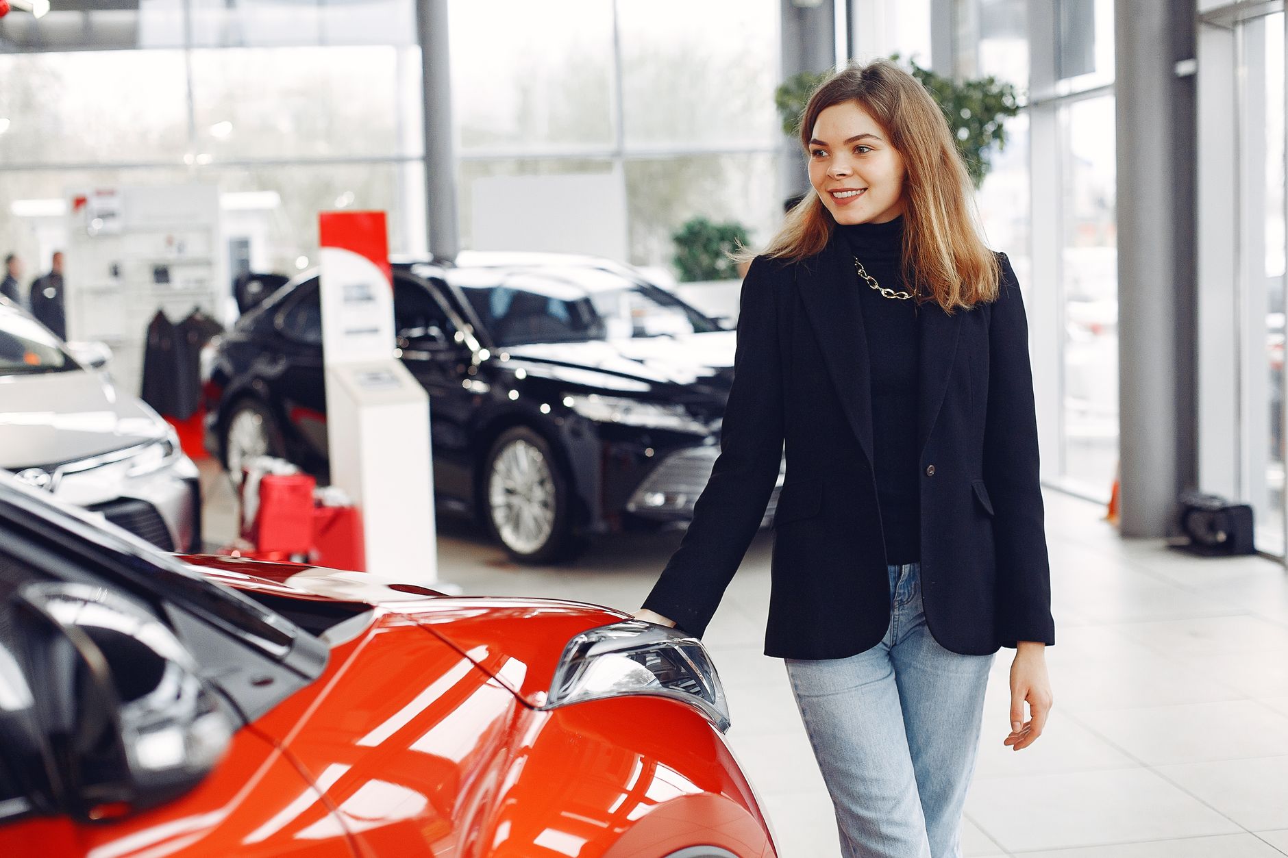 Happy stylish young female in jeans and black jacket standing near modern red shiny car in contemporary car showroom and looking at purchase