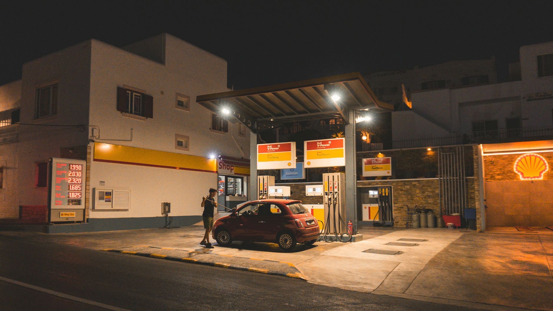 A person refuels a red car at a gas station at night, illuminated signs and lights.