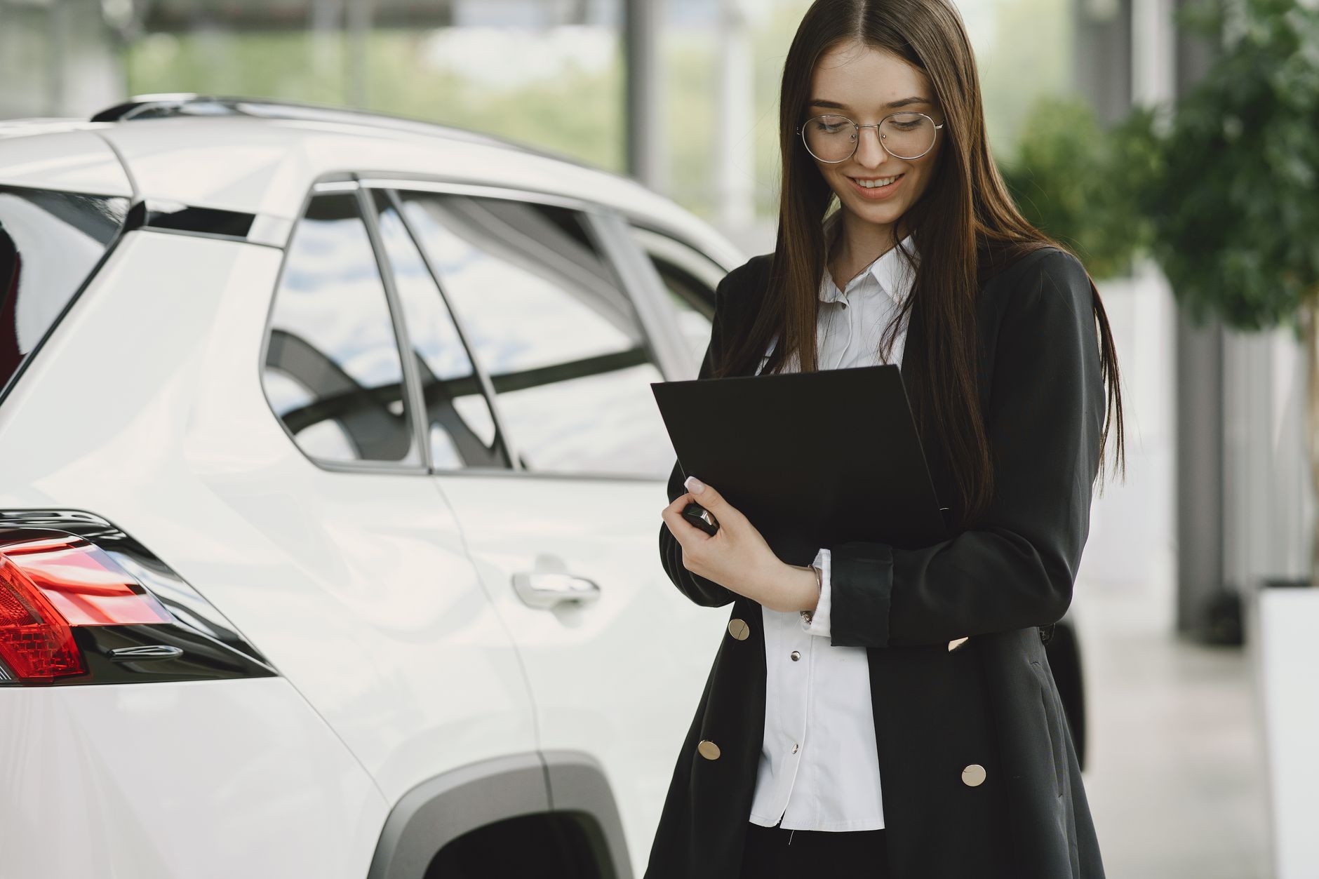 Smiling woman in a black jacket reviewing documents near a white car indoors.